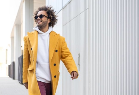 An afro man in a mustard coat, white hoodie, and sunglasses walks confidently along a modern, urban