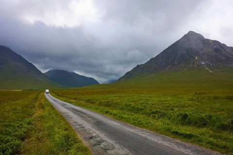 A lonely road leads through a vast landscape of dramatic mountain silhouettes and clouds, Glen