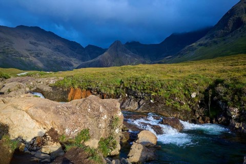 River flowing through a green landscape surrounded by mountains under a cloudy sky, Fairy Pools,