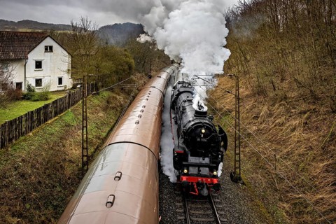 A steaming locomotive travels along a house and forest on winding tracks, historic steam locomotive