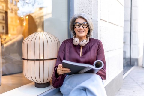 Mature woman sitting casually on a window ledge, smiling warmly at the camera while holding an open