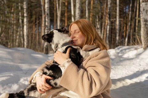 A woman warmly hugs her black and white Border Collie in a snowy forest, displaying affection