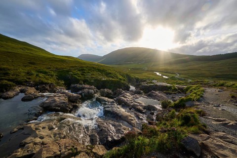 River flowing through rolling landscape in the warm light of sunset under cloudy sky, Fairy Pools,