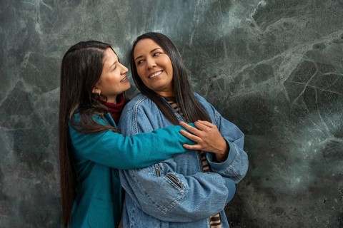 A joyful Latin lesbian couple shares a warm embrace outdoors, smiling lovingly at each other. The