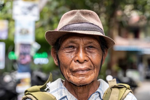An elderly balinese man wearing a hat and backpack, standing outdoors. The background is blurred,