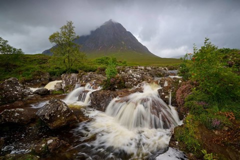 Impressive waterfall flows over rocks with a majestic mountain behind it under a cloudy sky,