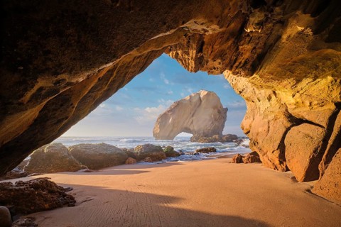 Discover the stunning Penedo do Guincho rock arch framed by a cave, with ocean waves and sandy