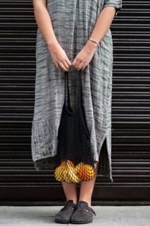 A woman in a long, gray dress carries a reusable mesh bag filled with bananas and oranges. The