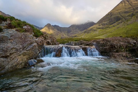 Waterfall with adjacent rocks in a green mountain landscape under cloudy sky, Fairy Pools, Grula,
