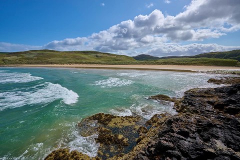 Waves crash against rocky coast against sandy beach and green hills under cloudy sky, Farr Bay,