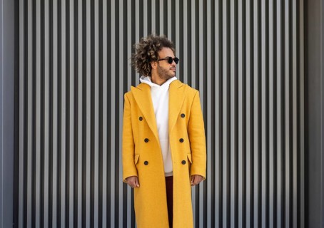 A stylish afro man in a mustard yellow coat stands confidently in front of a modern striped