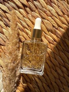 A luxurious glass cosmetic bottle containing gold infused oil rests beside dried grass on a woven