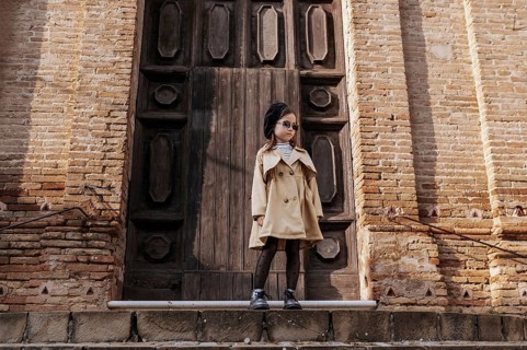 A stylish young girl in a trench coat and sunglasses stands confidently by a historic wooden