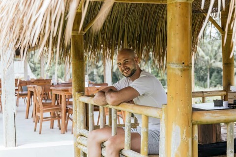 A man sits comfortably in a bamboo hut, surrounded by wooden furniture and lush greenery in Bali.