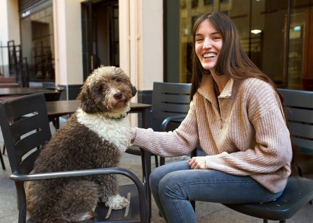 A cheerful young woman sits outdoors, beaming while gently petting her fluffy dog seated beside