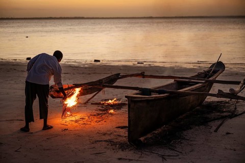 Llocal man use a small fire on a sandy beach at twilight to repair or seal a traditional wooden