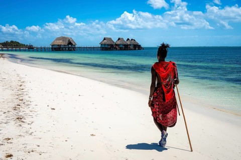 A Maasai man in traditional red attire walks away from the camera along a pristine white sand beach