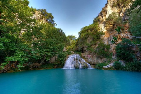 A waterfall plunges into a large turquoise-blue pool surrounded by green foliage and rocks,