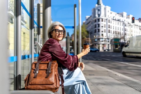 Senior woman with gray hair and glasses sitting at a city bus stop on a sunny day, holding a
