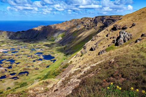 Rocky volcanic slopes overlooking a crater with water and the ocean behind it, the Ranu Kau volcano