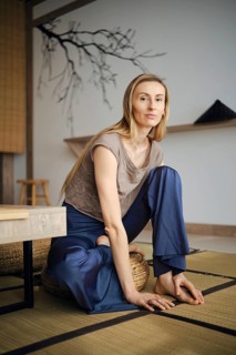 A woman sits on a mat in a room with minimal decor. She has long hair and wears comfortable clothes