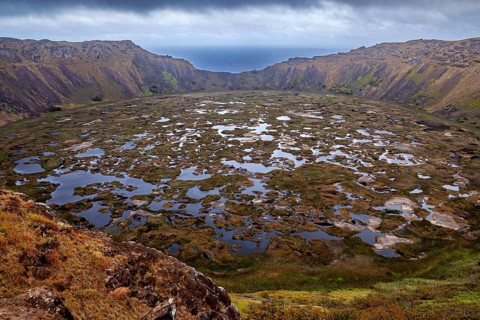Large crater with expanses of water surrounded by rocky and grassy walls under cloudy skies, the
