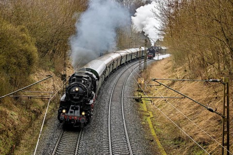 A steaming locomotive pulls a train through a wooded and winding route, historic steam locomotive