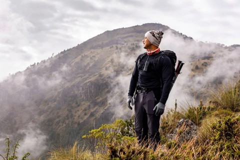 A hiker stands on a misty mountain slope, gazing into the cloudy horizon. Dressed warmly and