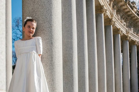 A young girl in a white First Communion dress stands by majestic stone columns. The sun illuminates