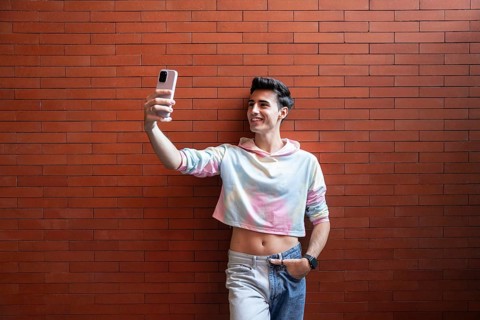 A young man smiles while taking a selfie with his smartphone against a vibrant red brick wall. He