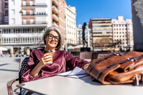 Mature woman with gray hair and glasses smiling while enjoying a takeaway coffee at an outdoor