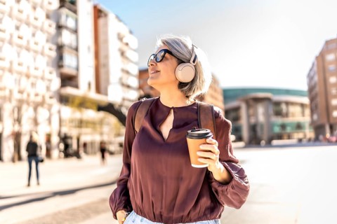 Mature woman with gray hair wearing sunglasses and headphones, smiling while enjoying a hot coffee