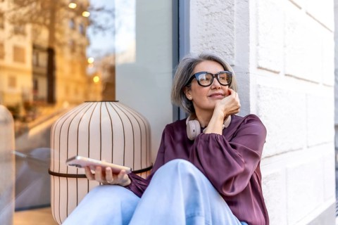 Grey haired woman with headphones around her neck, smiling thoughtfully on an urban sidewalk while