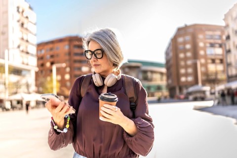 Senior woman walking in a city plaza, looking at her smartphone while holding a takeaway coffee and