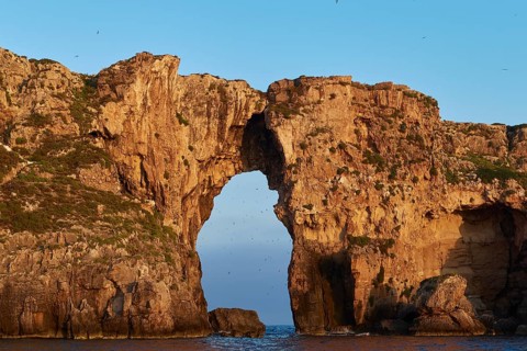 An imposing rock gate stretches across the calm sea under clear skies, morning light, Tsichli Babas