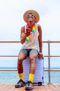 An LGBTQIA+ man dressed in vibrant attire, seated on a suitcase by the sea, joyfully preparing for