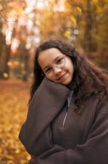 A teenage girl in glasses and warm sweater relaxes amidst vibrant autumn foliage in a forest. The