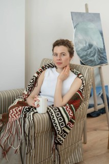 A woman sits comfortably in a armchair, wrapped in a patterned blanket, holding a coffee mug.