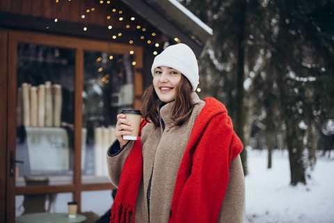 A cheerful young woman, standing outside a cozy cafe, enjoys a warm beverage on a cold winter day.