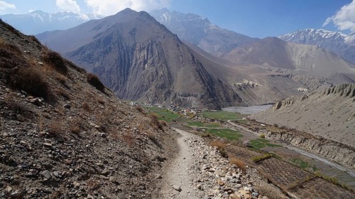 Gravelly mountain trail with views of fields and surrounding mountains under a blue sky, trekking