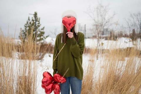 Woman in green sweater and winter hat holding heart shaped balloons in a snowy park. The serene