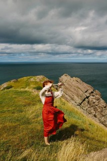 A woman in a vintage red dress and sunglasses using a spyglass on a windy coastal cliff at Middle
