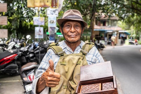 A smiling balinese vendor in casual attire and a hat gives a thumbs up while showcasing handcrafted