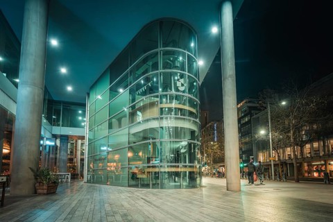 A modern glass-fronted building is illuminated at night as people pass by, New Year's Eve London,