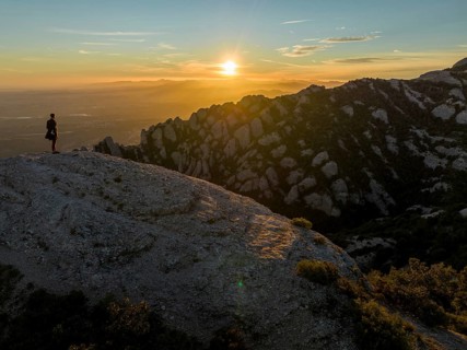 A lone figure stands on a rocky cliff at Montserrat, Catalonia, as the sun rises, casting warm