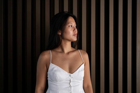A young Chinese woman in a stylish white dress stands confidently against a wooden backdrop. The