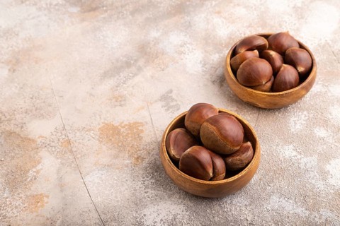 Wooden bowl with raw edible ?hestnuts on brown concrete background, side view, copy space,