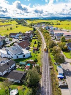 Yellow train travelling through a village surrounded by a rural landscape and houses, tamping