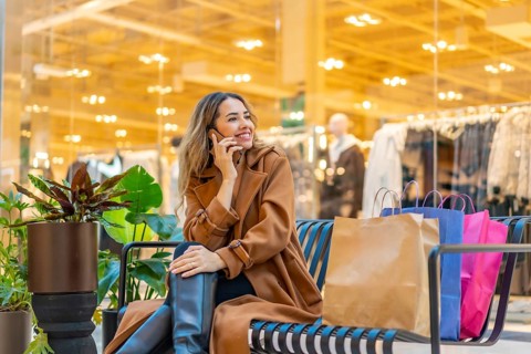Young woman sits on a bench in a modern mall, smiling and talking on her smartphone with shopping