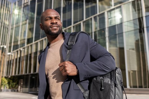 A confident cuban man in a blazer and sweater, carrying a backpack, stands outside a modern office